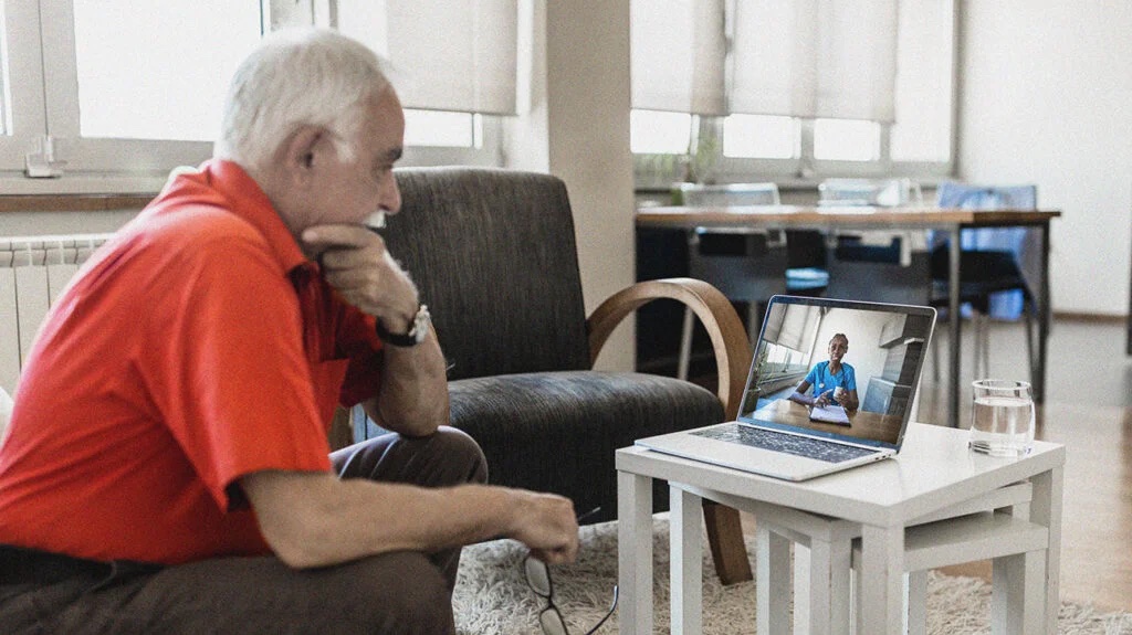 An elderly man sitting on a couch, engaged in telethrapy through an video call on his laptop.