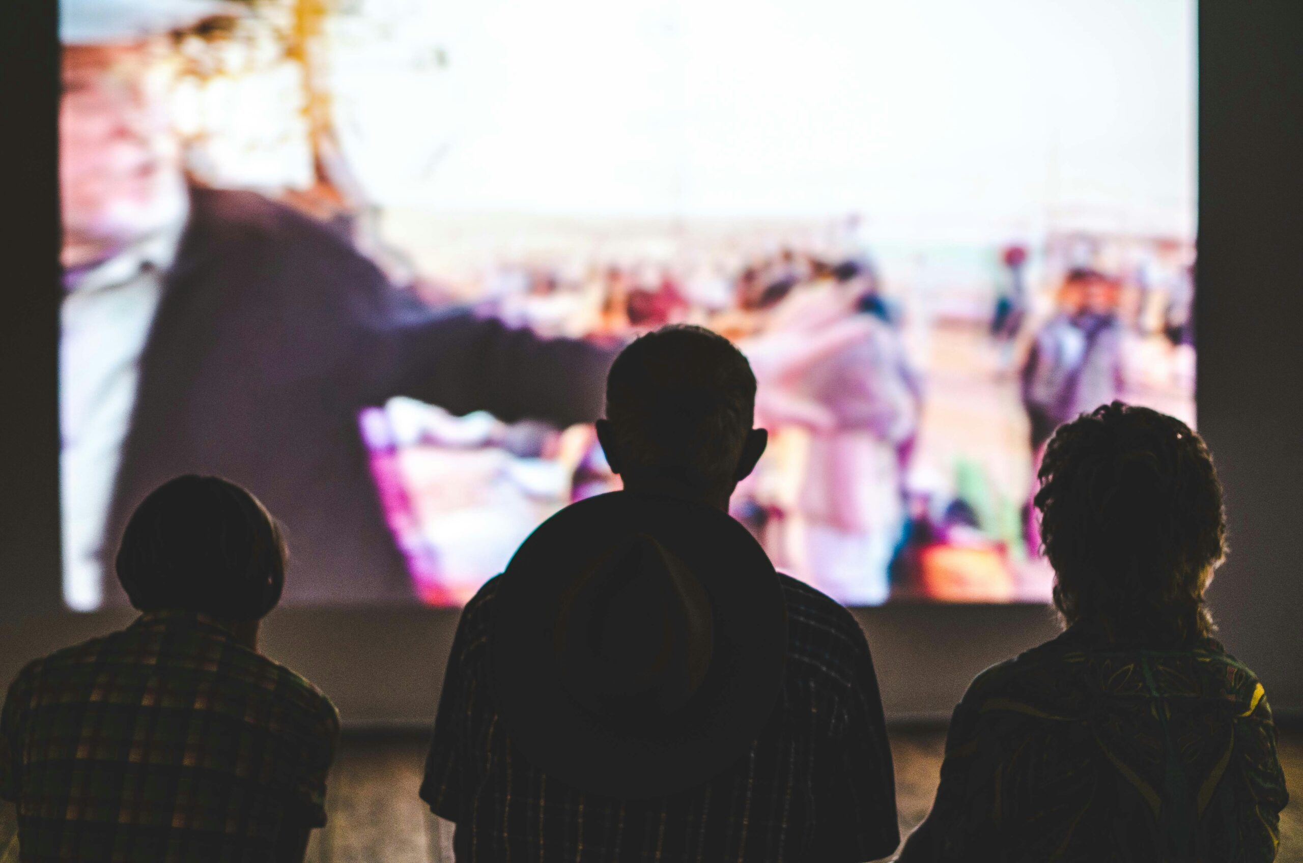 Silhouette of 3 people watching a movie on a large screen.