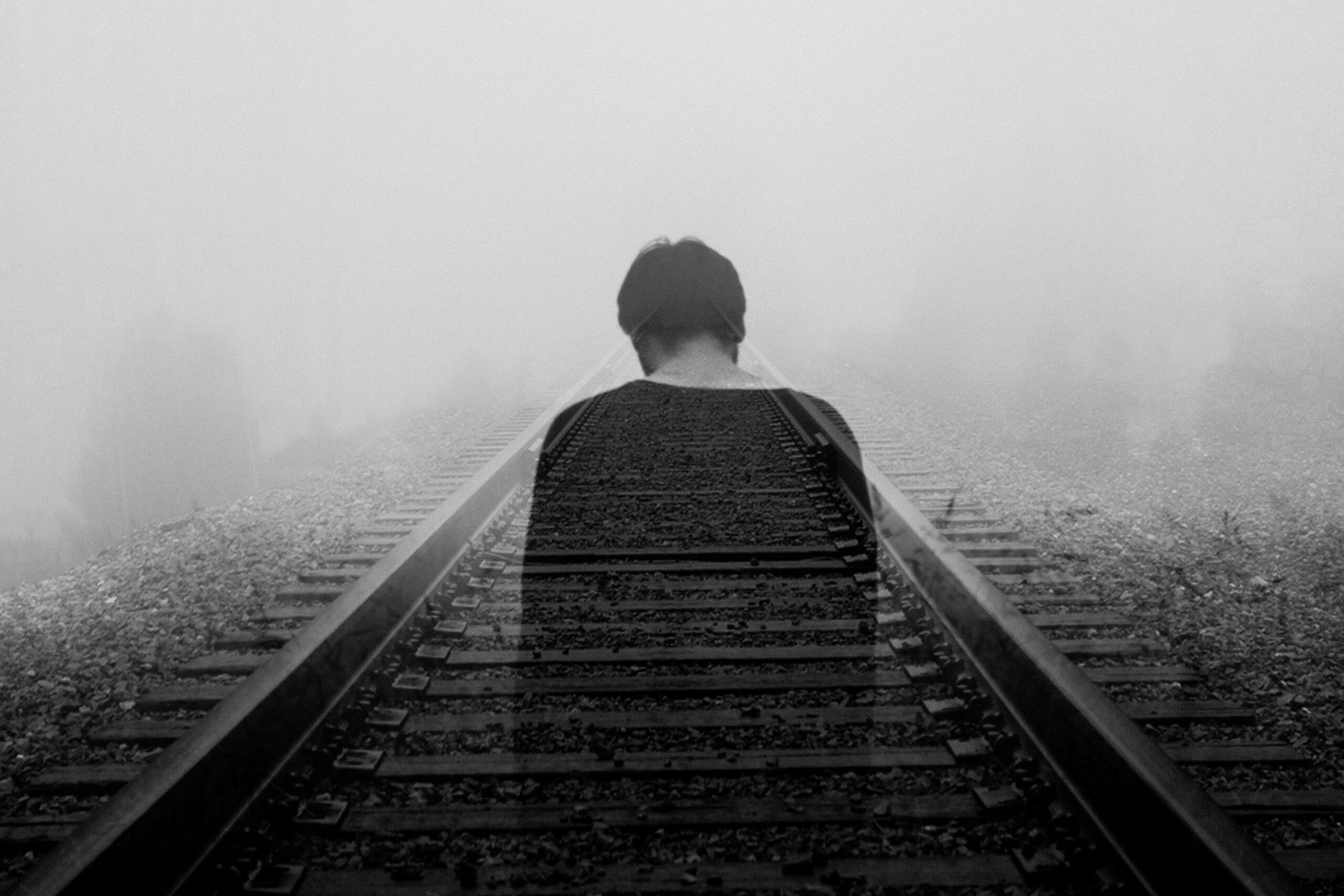 Grayscale photo of a man wearing a black shirt, standing on the railway tracks.