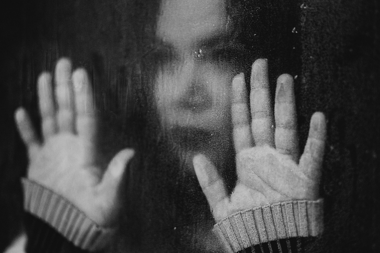 Hands up against a window in the foreground, a woman, in soft focus, faces the window with a dead-eyed expression