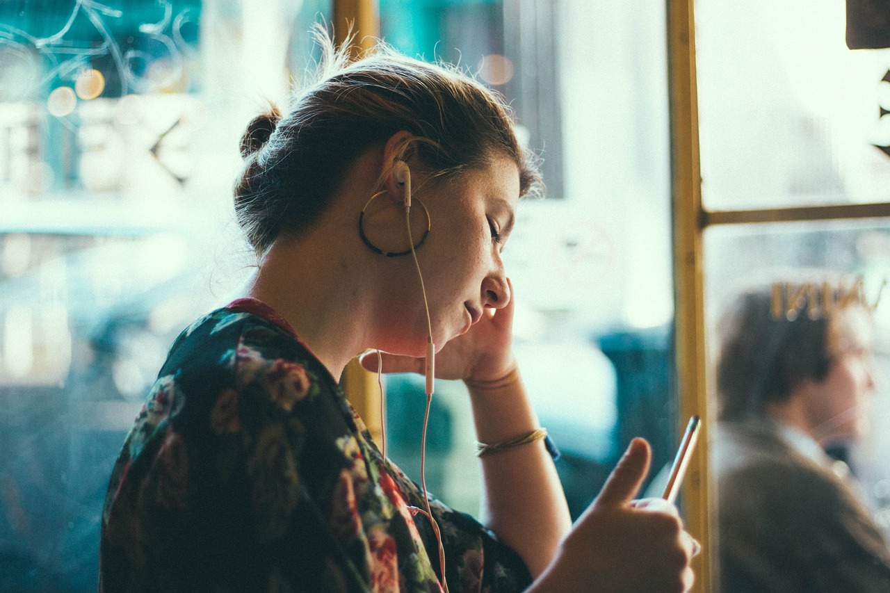 Woman seated next to a window, earphones plugged in, looking down at something with a pen in hand
